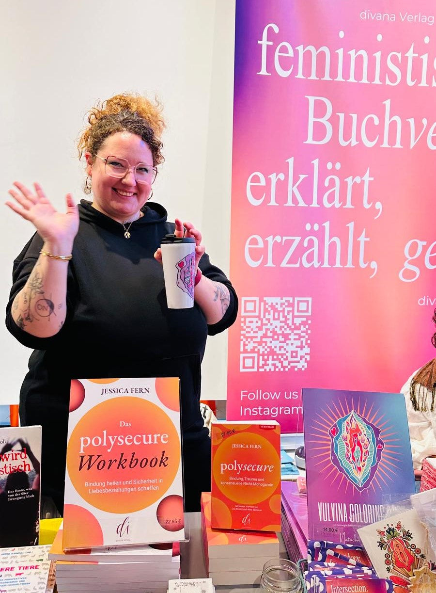 Auf dem Bild sieht man Natacha Jill Colin, wie sie auf der feministischen Buchmesse in Berlin den Stand des divana Verlags betreut. Hier liegen unter anderem das Buch Polysecure, Vulvina Malbuch, It's all blood und das Polysecure Workbook aus.